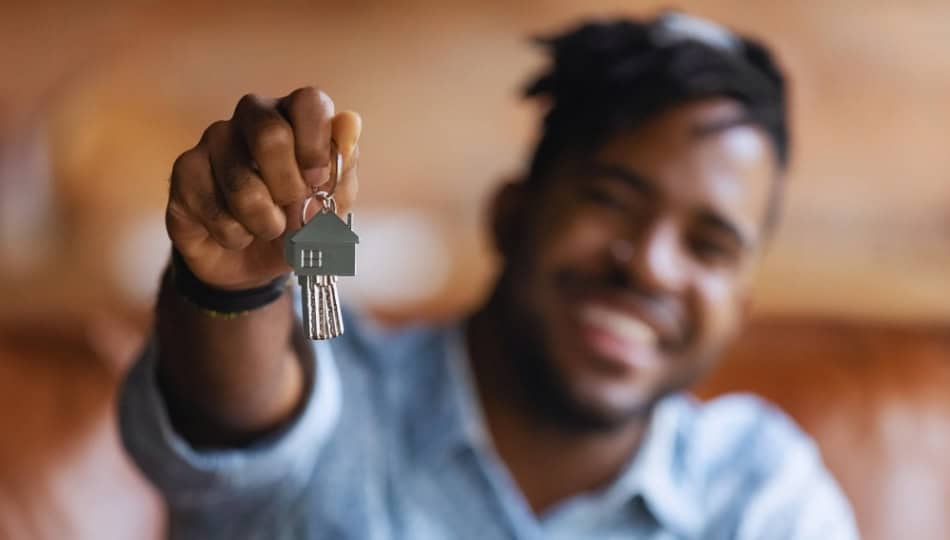 A person holding up a keychain with a house-shaped pendant and keys, smiling in the background. The image is focused on the keys, with the person slightly out of focus.
