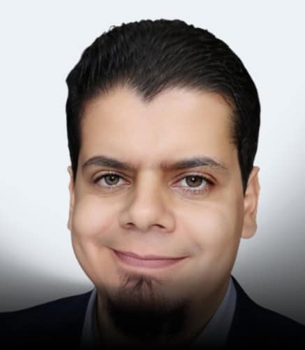 A man with short, dark, slicked-back hair and facial hair is smiling slightly. He is wearing a dark suit jacket and looking directly at the camera against a light, plain background.