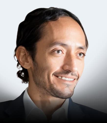 A man with medium-length dark hair and facial hair is smiling. He is wearing a white collared shirt and a dark blazer. The background is a light, plain color.