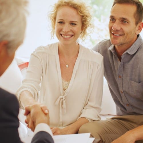 A woman and a man sit closely together, smiling, as the woman shakes hands with another person across a table. Documents are visible in the foreground.