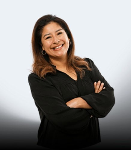 A woman with medium-length brown hair stands smiling with her arms crossed, dressed in a black long-sleeve blouse, in front of a light grey background.