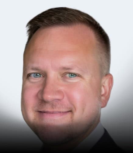 A man with short light brown hair, blue eyes, and fair skin is smiling. He is wearing a dark suit jacket and white shirt. The bottom of the image is darkened. The background is light gray.