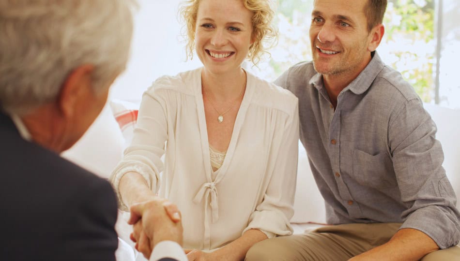 A woman and a man sit closely together on a couch, smiling as the woman shakes hands with an older man across from them. The setting appears to be a casual meeting or consultation.