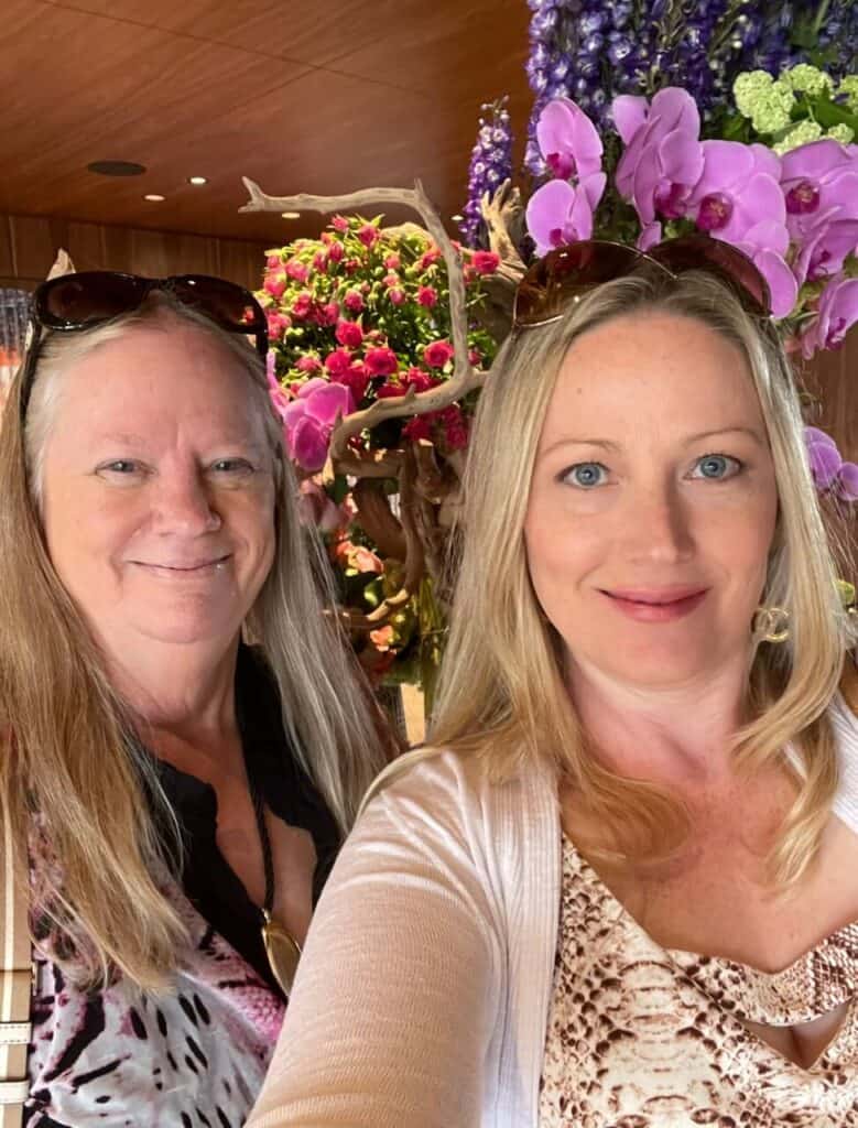 Two women standing indoors, smiling at the camera. There are large arrangements of pink and purple flowers in the background. Both women have long, light hair and are wearing sunglasses on their heads.