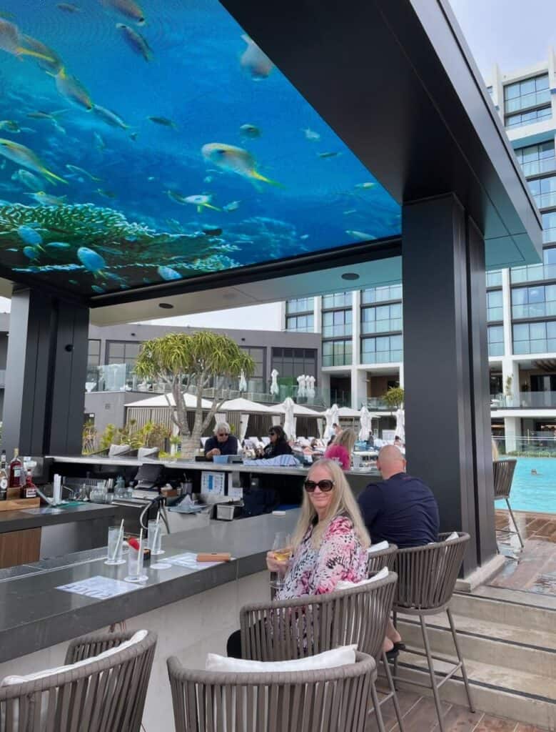 A woman wearing sunglasses sits at an outdoor bar with a drink in hand. Behind her, people lounge by a pool. An overhead panel displays an underwater scene with fish. Modern buildings surround the area.