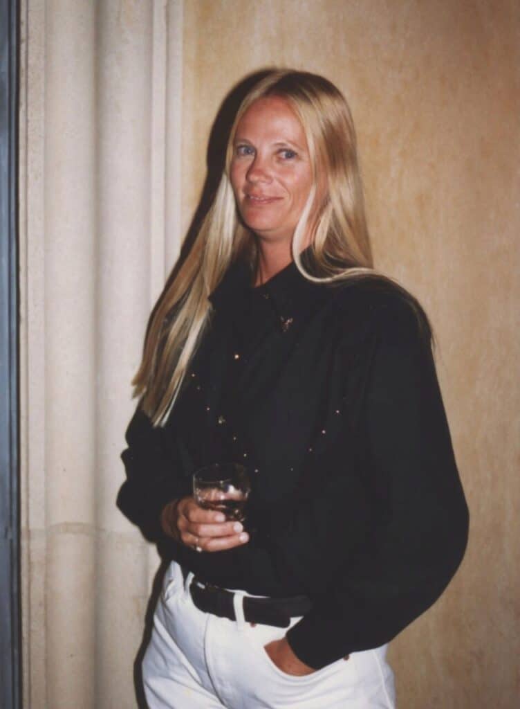 A woman with long blonde hair stands indoors against a beige wall, wearing a black shirt and white pants. She is holding a glass in her right hand and looking at the camera with a slight smile.