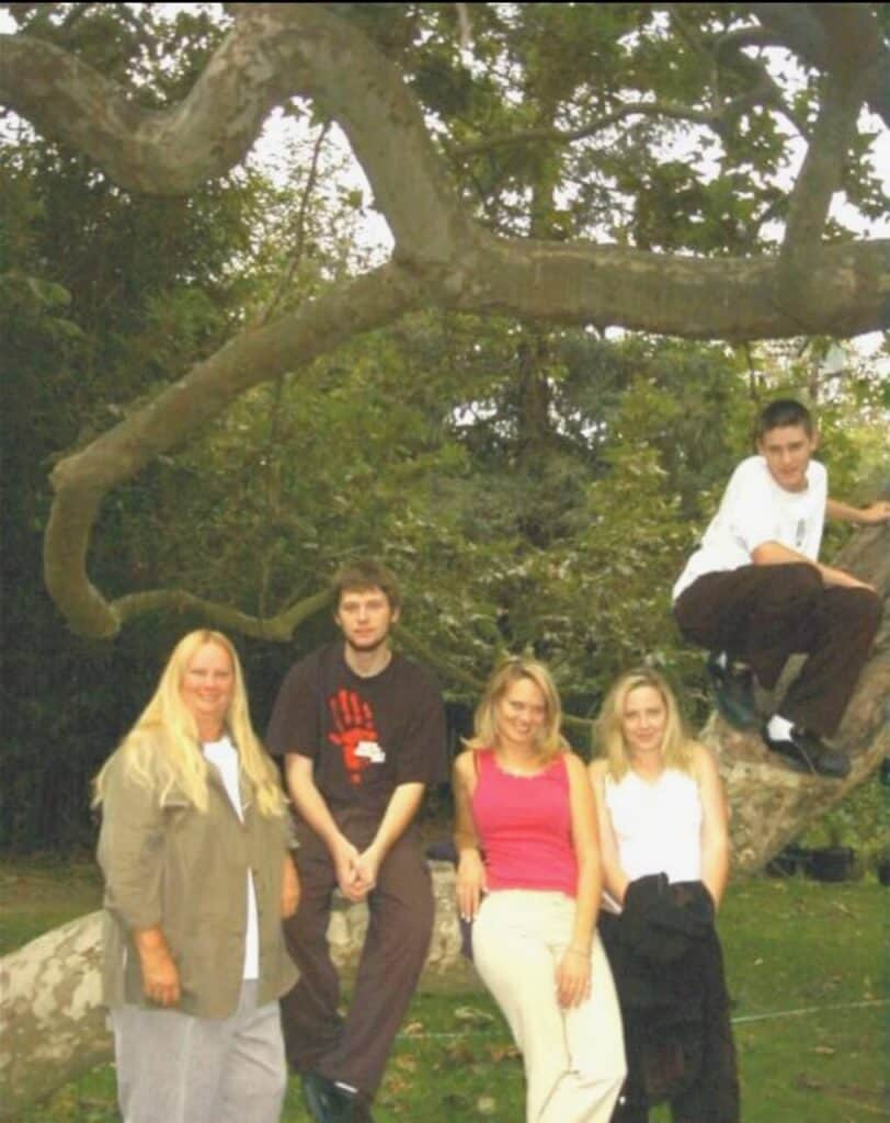 Five people pose outdoors near a large, curved tree branch. Three are sitting or leaning on the branch, while two stand in front of it. The group is surrounded by greenery and trees.