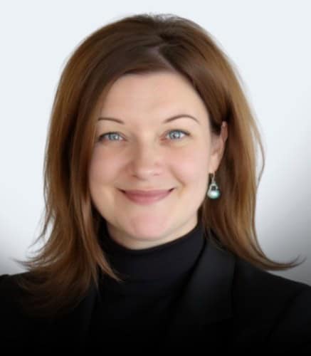 A woman with shoulder-length brown hair wearing a black turtleneck and a black blazer, smiling at the camera. She has blue eyes and is wearing a single dangling earring. The background is plain and light-colored.