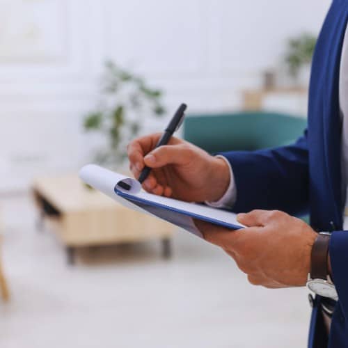 A person in a blue suit is holding a clipboard and writing with a pen. The background is blurred and shows a light-colored room with plants and furniture.