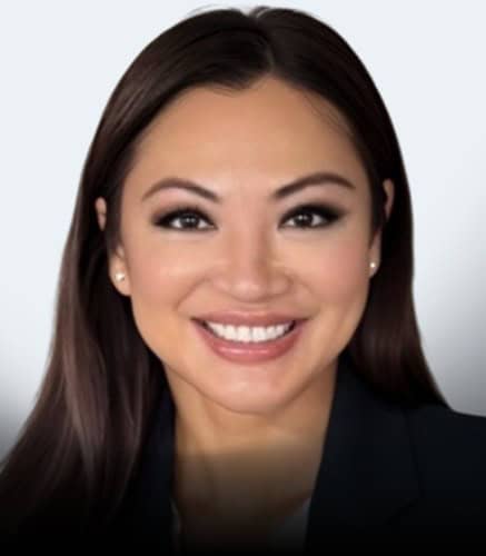 A woman with long straight brown hair, wearing a dark blazer and small stud earrings, smiles at the camera against a plain light gray background.