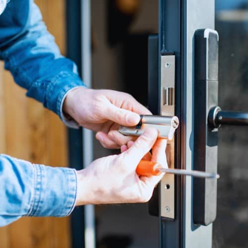 A person in a denim shirt uses a screwdriver to install or remove a cylinder lock from a metal door.