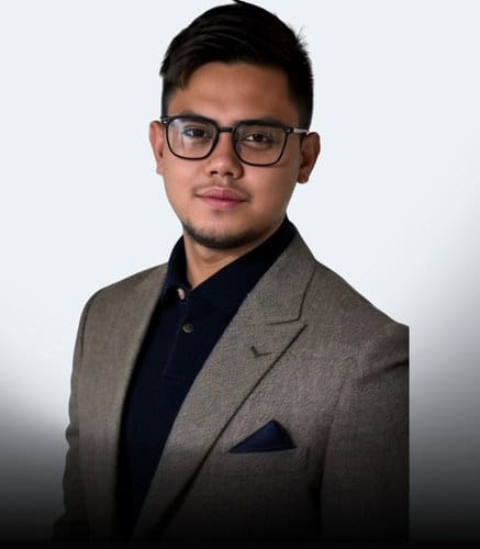 A man with short dark hair and glasses is wearing a brown blazer over a black collared shirt. He faces forward with a neutral expression against a plain light background.