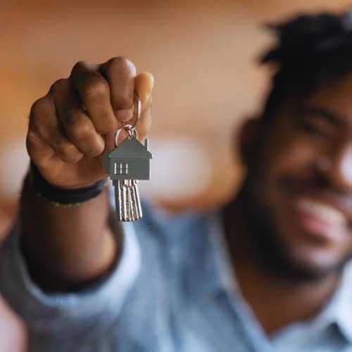 A person holds out a set of keys on a house-shaped keychain, with the focus on the keys. The individual is smiling in the blurred background.