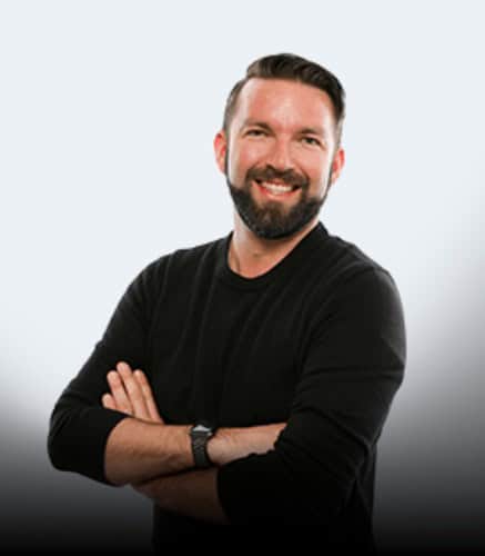 A man with short dark hair and a beard is smiling with arms crossed, wearing a black long-sleeve shirt and a watch, standing against a neutral gradient background.