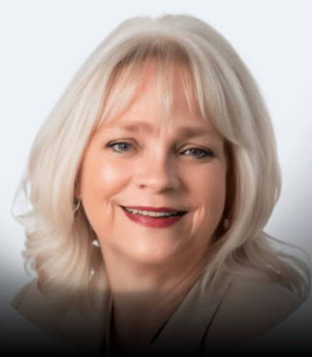 A woman with shoulder-length white hair and light skin is smiling. She is wearing a beige blazer and has red lipstick. The background is plain and light-colored.