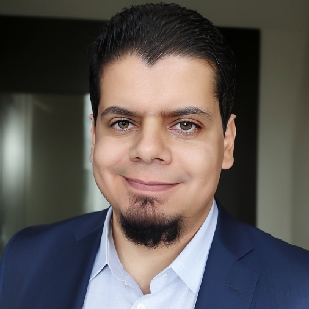 A man with dark hair and a short goatee is wearing a navy blue suit jacket and a light blue shirt, smiling at the camera. He is posed indoors against a blurred background.