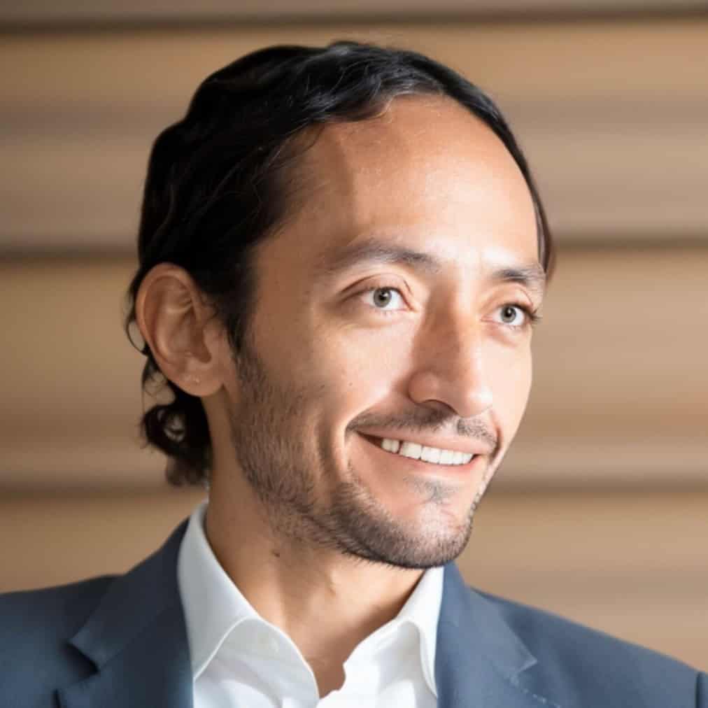 A man with wavy dark hair and a trimmed beard is wearing a white shirt and a dark blazer, smiling and looking slightly to the side. The background features horizontal blurred lines in beige tones.