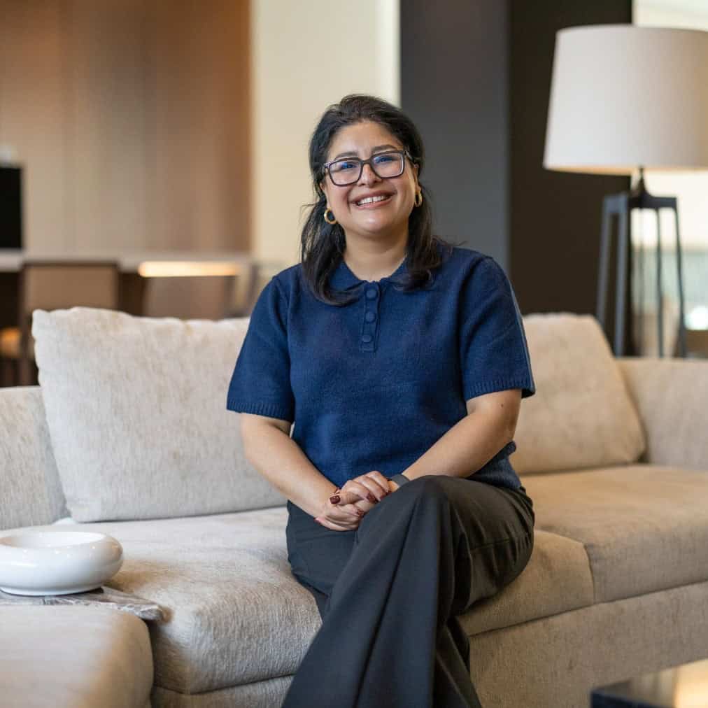 A woman with glasses and dark hair sits on a light-colored sofa in a modern living room, smiling at the camera. She is wearing a navy blue top and dark pants, with her hands folded in her lap.