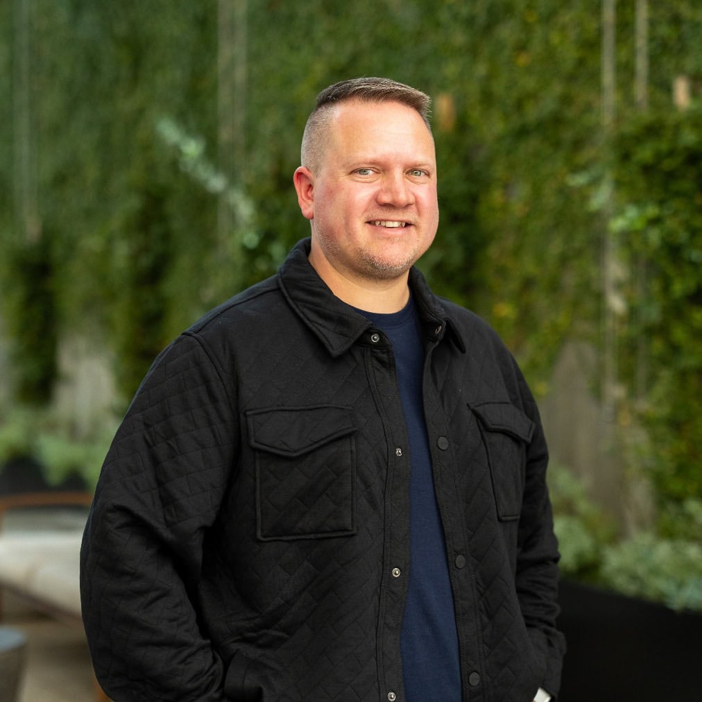 A man wearing a black quilted jacket and a dark shirt stands outdoors in front of a green, leafy background, looking at the camera and smiling.