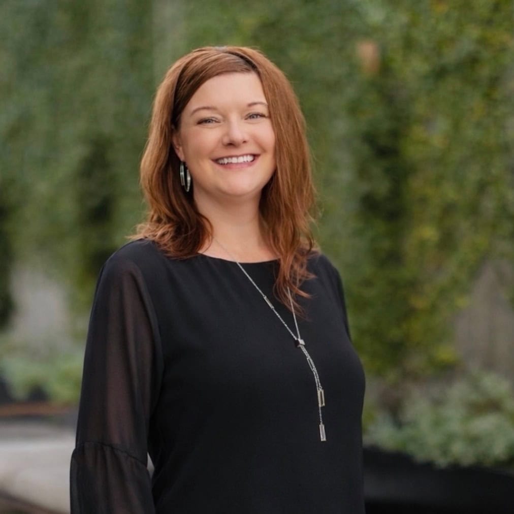 A woman with medium-length reddish-brown hair wearing a black blouse and a long silver necklace is smiling outdoors. The background features greenery and blurred foliage.