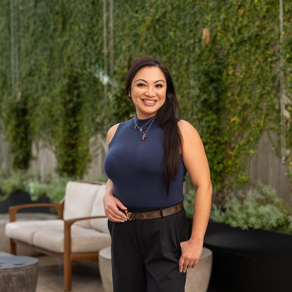 A woman with long brown hair stands outdoors, smiling at the camera. She wears a sleeveless navy top, black pants, necklaces, and a belt. Behind her are a beige cushioned chair and green plants on a vertical garden wall.