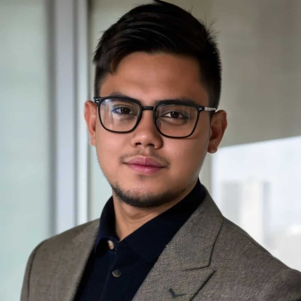 A man with short dark hair, glasses, and trimmed facial hair is wearing a brown blazer over a black shirt. He is posing indoors near a window with a blurred cityscape in the background.
