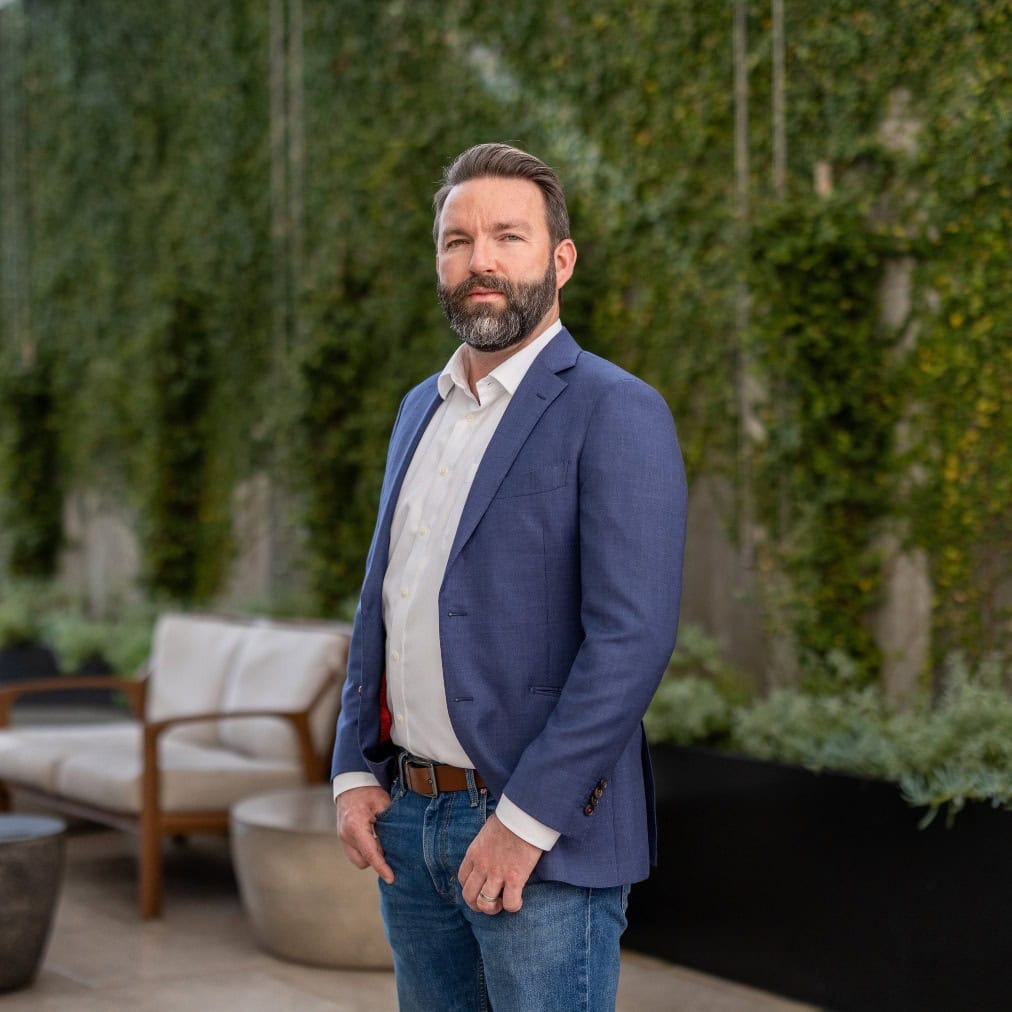 A man with a beard wearing a blue blazer, white shirt, and jeans stands outdoors in front of a wall covered with green vines, with outdoor seating and planters visible behind him.
