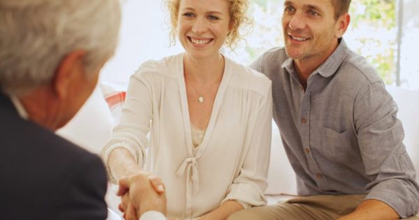 A woman and a man sit closely together on a couch, smiling as the woman shakes hands with an older man across from them. The setting appears to be a casual meeting or consultation.