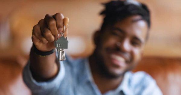 A person holding up a keychain with a house-shaped pendant and keys, smiling in the background. The image is focused on the keys, with the person slightly out of focus.